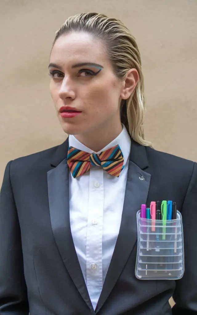 A photo of a quirky woman dressed in a smart casual outfit. She is wearing a pocket protector filled with colorful pens and a stylish bowtie. The background is a beige wall.