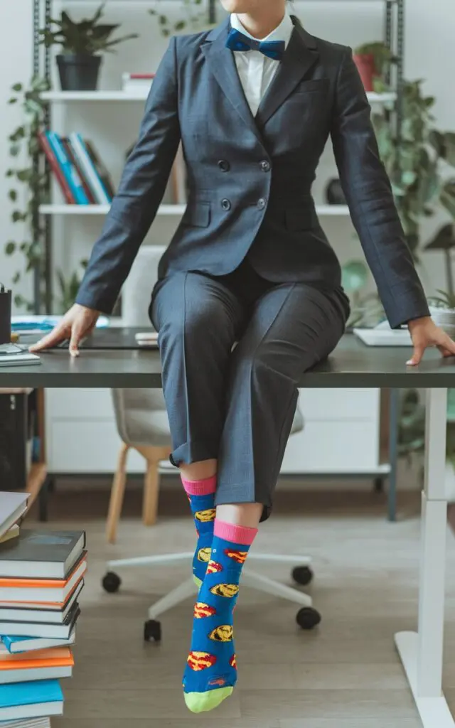A photo of a stylish woman in a tailored suit, sitting confidently on a desk with her legs crossed. She reveals brightly colored superhero socks. The desk is clean and there are books near it. The background is a room with shelves and plants.