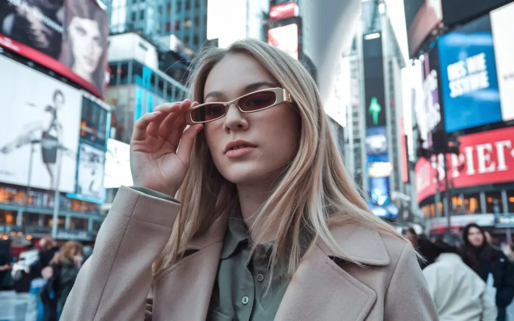 A photo of a young American woman wearing tiny elongated minimalist rectangular sunglasses in Times Square. She has blonde hair and is wearing a beige coat and a green shirt. The background is filled with billboards and bright lights.