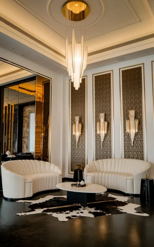A photo of an art deco luxurious modern living room with sculptural lighting. The room has a high ceiling, white walls, and a dark floor. There are two white sofas with curved lines, a black and white cowhide rug, a white coffee table, and a black side table. There is a gold pendent light fixture hanging from the ceiling. The walls have gold accents and a patterned texture.