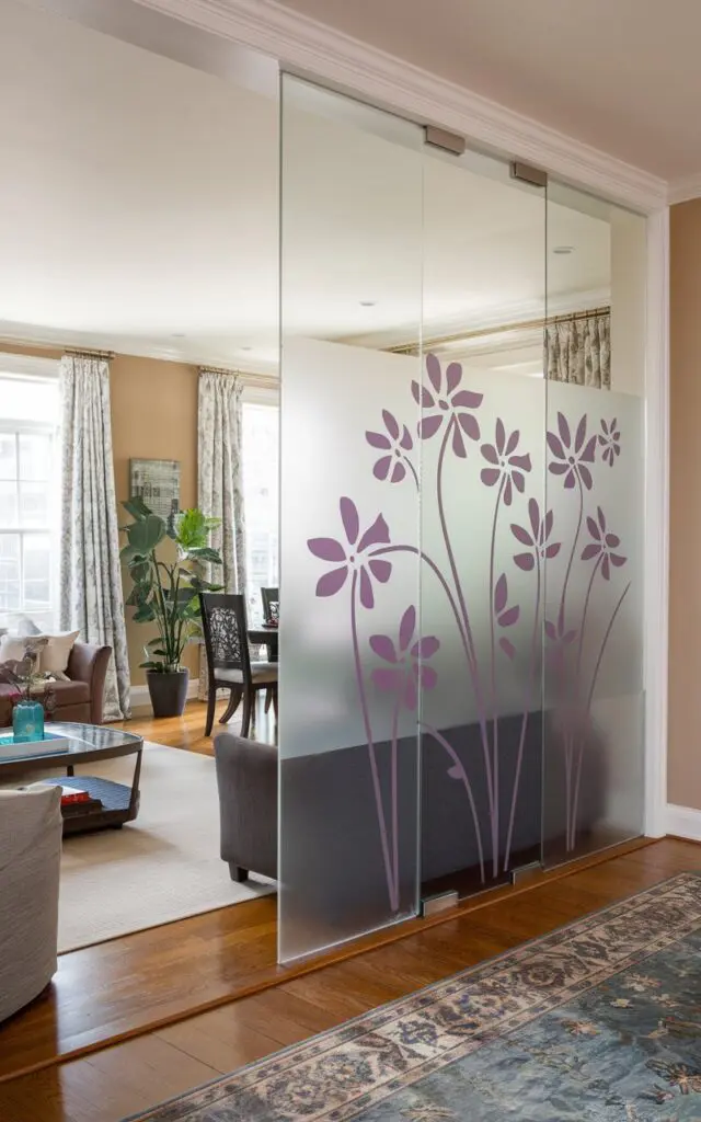 A photo of a transitional living room with a frosted glass partition with a purple floral design. The partition separates the living room from the dining area. The living room has a sofa, a coffee table, and a plant. The dining area has a dining table and chairs. The flooring is hardwood. The walls are painted beige.