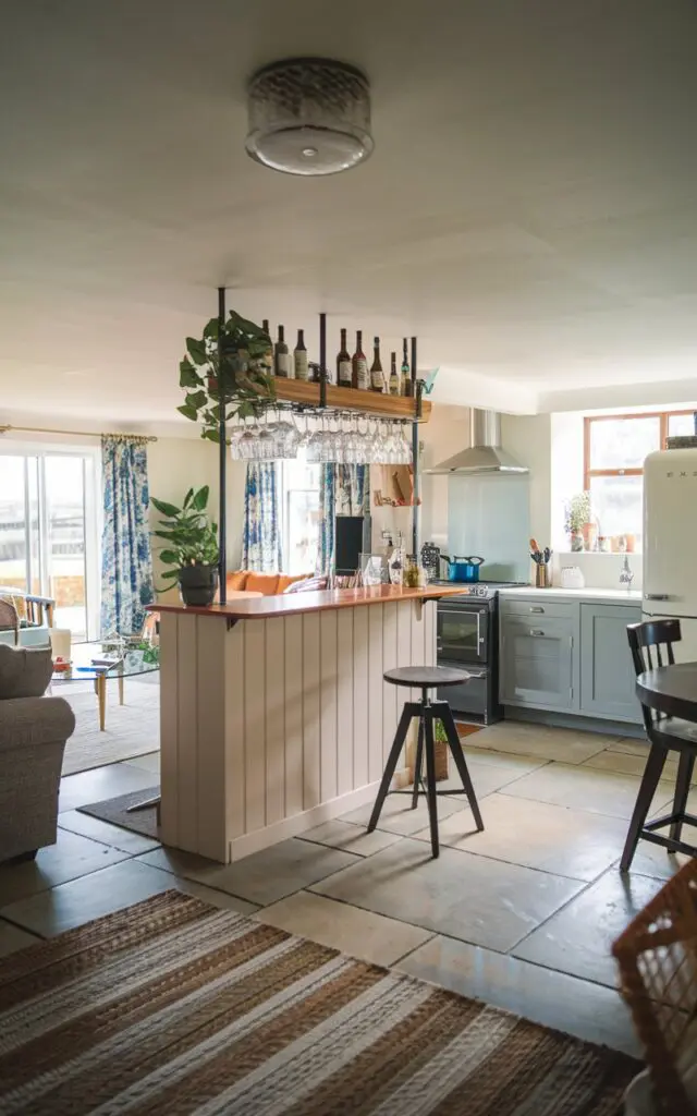 A photo of a modern English countryside home with a room divider featuring a stylish built-in bar. The bar is placed between the living room and kitchen. The living room has a sofa, a coffee table, and a plant. The kitchen has a stove, a refrigerator, and a sink. The ceiling has a light fixture. The floor is tiled and has a rug. The light is diffused.