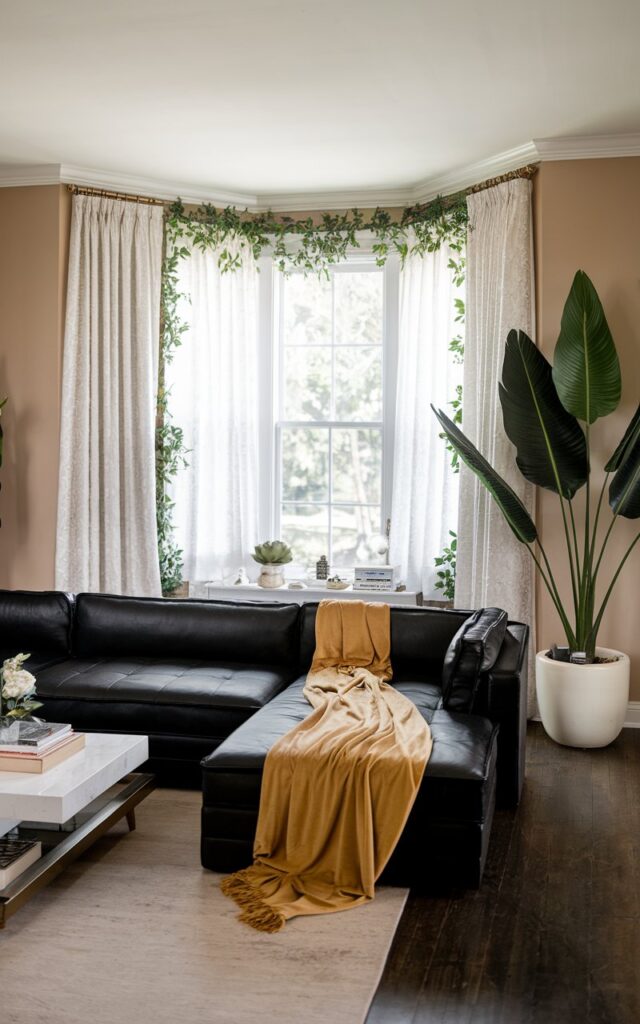 A photo of a Hollywood Regency living room with a gold throw blanket on a black sectional sofa. The room has a beige wall, a white coffee table, and a few pieces of decor. There is a large potted plant near the window. The window has white curtains and is surrounded by greenery. The flooring is a dark wood.