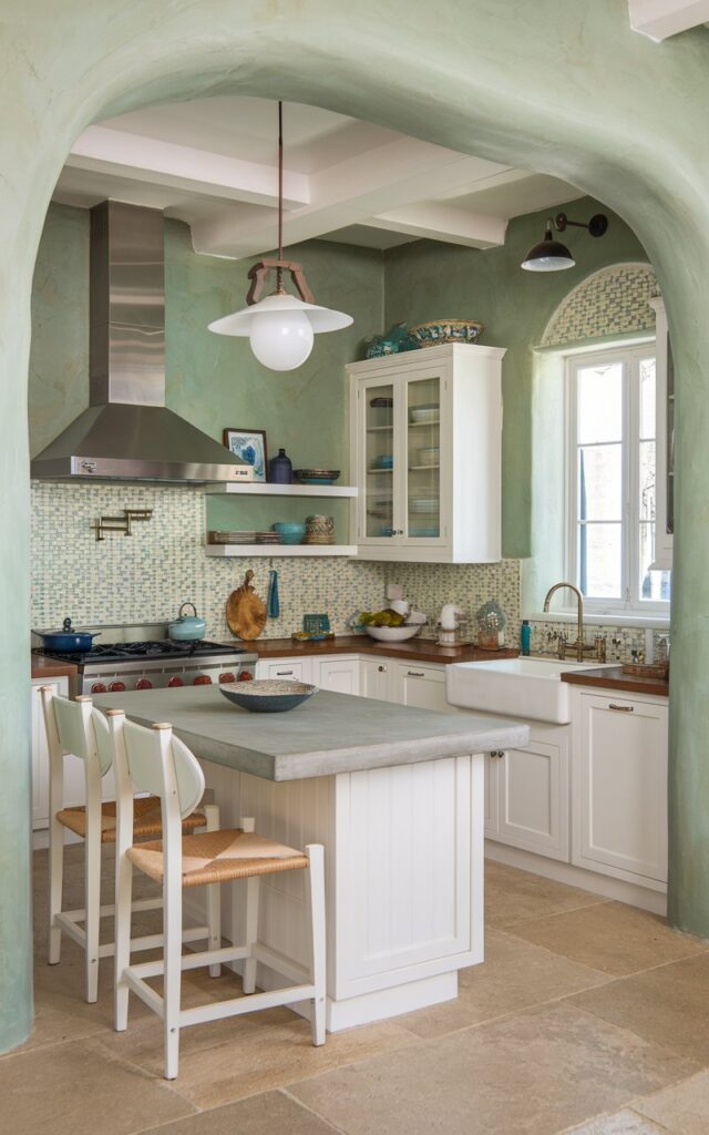 A photo of a California coastal-style kitchen with sage green textured walls and white cabinets. The kitchen is fully furnished with a modern pendant light hanging above a white island with a concrete countertop. There's a stainless steel range hood above the stove. The backsplash is a mosaic of blue and white tiles. There are white chairs under the window. The floor is made of large beige tiles.