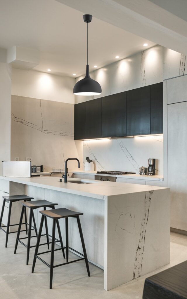 A photo of a monochromatic modern transitional kitchen with a white, black, and grey color scheme. The kitchen island has a matte finish and features veining in the stone countertops and walls. The kitchen is fully furnished with stools and appliances. A stylish hanging black pendant light with a light on is suspended from the ceiling. The lighting in the kitchen is soft and diffused throughout.