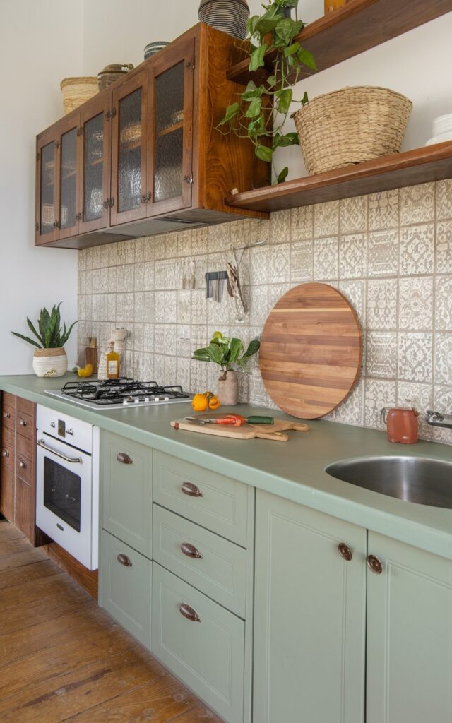 A photo of a fully furnished Balinese farmhouse kitchen with a matte sage green countertop. The kitchen features wooden cabinets, a white oven, and a stainless steel sink. There's a large wooden cutting board on the counter, along with a knife and a few vegetables. A wicker basket and a potted plant are also on the counter. The backsplash has a beige and grey patterned tile. The floor is made of wood.