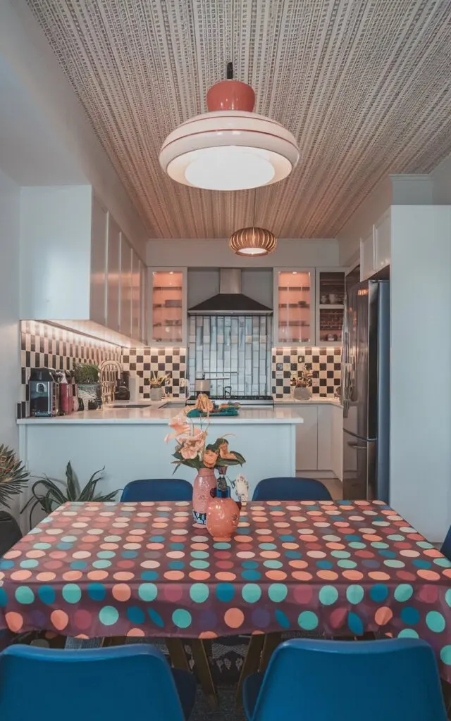 A photo of a fully-furnished modern Balinese kitschy kitchen with a retro polka dot tablecloth on the dining table. The ceiling has retro wallpaper and a retro pendant light. There is a vase of flowers on the table. The kitchen has a smoothie maker, a blender, and a toaster. The floor is made of wood.