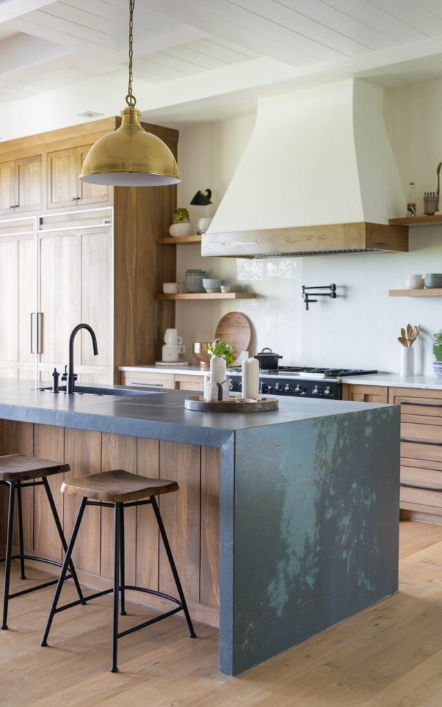 A photo of a modern farmhouse kitchen with a sleek island. The island has a waterfall countertop made of grayish-blue stone. There are two bar stools near the island. A golden pendant light hangs above the island. The kitchen has wooden cabinets, a white range hood, and a black sink.