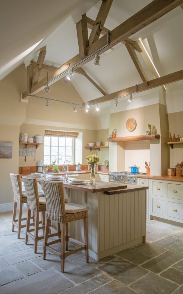 A photo of a fully-furnished English countryside kitchen with a kitchen island. There is a breakfast bar at the island. The kitchen has a vaulted ceiling with adjustable track lighting. The lighting illuminates the kitchen.