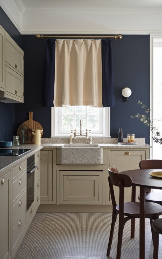 A photo of a Bauhaus-style kitchen with a beige and navy blue two-tone curtain on a small window above a granite drop-in sink. The curtain matches the rest of the kitchen's color scheme, which includes beige cabinets, navy blue walls, and a beige-and-grey checkerboard floor. The kitchen is fully furnished, with a wooden table, chairs, and a few decorative items. The natural light is filtered through a nearby window, casting a soft glow on the room.