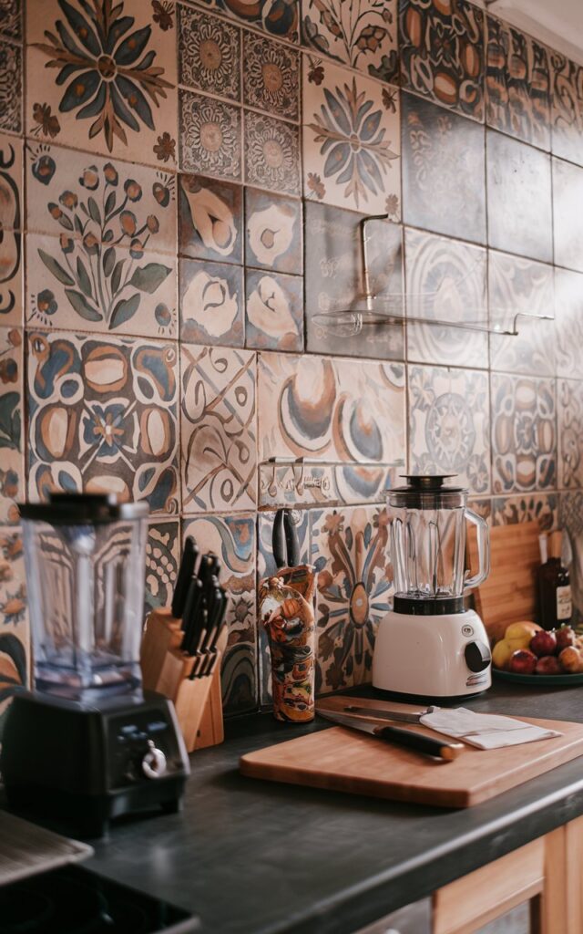A photo of a boho kitchen with a backsplash of hand-painted tiles in intricate floral designs and tribal-inspired art. The counter is fully furnished with various kitchen items, such as a blender, a knife, and some fruits. The natural light is soft and casts a warm glow on the tiles. The overall ambiance is rustic and cozy.