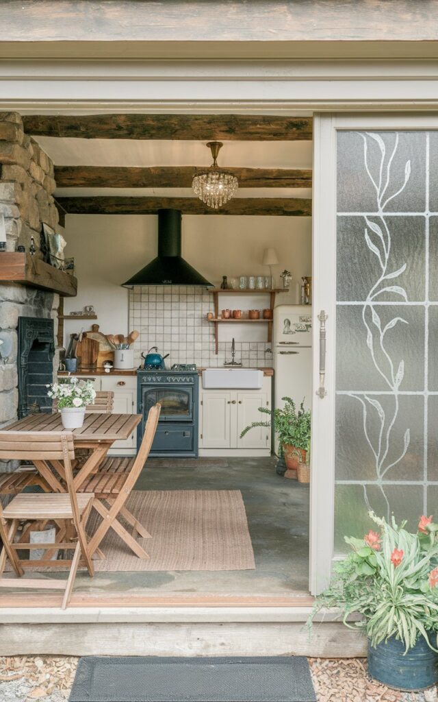 A photo of a fully-furnished English countryside open kitchen with an outdoor patio. The kitchen has a rustic charm with wooden beams, a stone fireplace, and a concrete floor. The kitchen is equipped with a stove, oven, fridge, and sink. There is a wooden table and chairs in the outdoor patio, with potted plants. A sliding door with a floral design in frosted glass connects the kitchen with the outdoor patio. The floor has a rug, and the ceiling is trimmed and has a crystal pendant light.