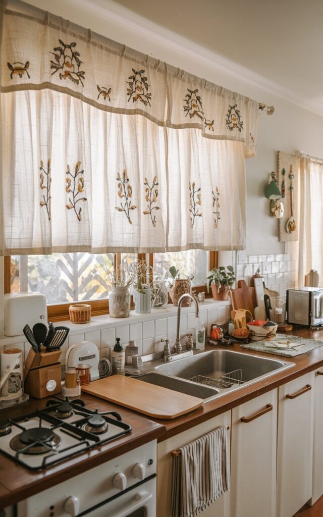A photo of a mid-century modern kitchen with an embroidered valance and a soft curtain on the window above the sink. The kitchen and counter are fully furnished with appliances, utensils, and decor. The embroidery on the valance is distinct and clear. The natural lighting is filtered through the window.