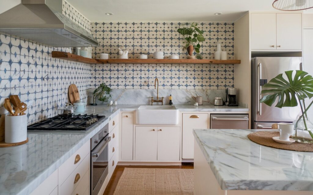 A photo of an L-shaped boho and coastal kitchen with a marble countertop and an unusual backsplash. The kitchen is fully furnished and contains essential items such as a stove, oven, sink, and refrigerator. There is a wooden shelf above the sink and a potted plant near the refrigerator. The backsplash features a pattern of blue and white tiles. The floor is covered with a beige rug.
