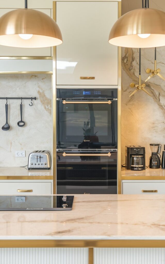 A photo of a modern glam kitchen with a luxurious countertop. There is a double oven with touch-screen control on the countertop. The oven has a black finish and is positioned between two gold pendant lights. The countertop has a marble texture and is gold-edged. There are other kitchen appliances, such as a toaster and a coffee maker, on the countertop. The background has a beige wall with a few hooks.