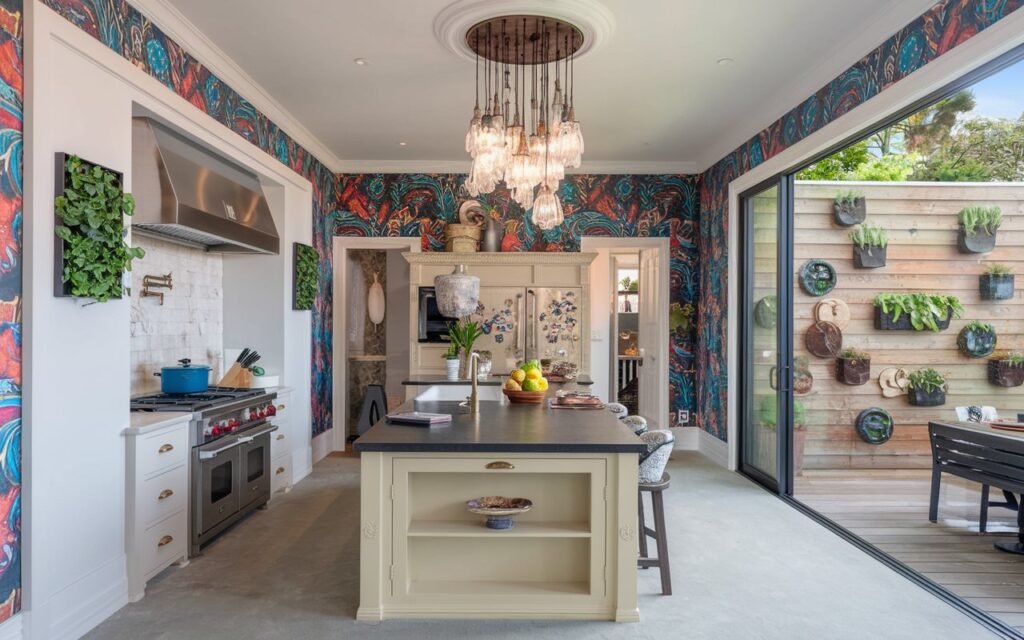 A photo of a modern and traditional kitchen with a touch of AI and vintage elements. The walls have bold, bright wallpaper. There's a cluster of crystal pendant lights above the island. The kitchen has a plant wall on one side of the wall. A sliding door leads to an outdoor patio for cooking. The kitchen and island are fully furnished. The natural light is soft and even.