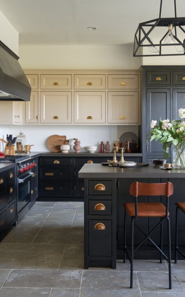A photo of a transitional kitchen with an L-shaped layout. The lower cabinets are dark in color, while the upper cabinets are light. There is a large island in the center of the room with seating. The cabinets have gold, matte black, and leather pulls. The floor is made of large concrete tiles.