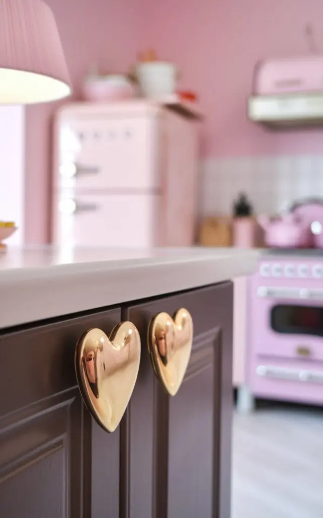 A photo of a fully furnished girly kitchen with cabinets having heart-shaped handles in ceramic gold. The photo is focused on a closer view of the heart-shaped handle. The handle is placed on a dark cabinet. The background is blurred, revealing a kitchen with pink walls, a pink refrigerator, and a pink stove. There is also a pink lampshade on the counter.