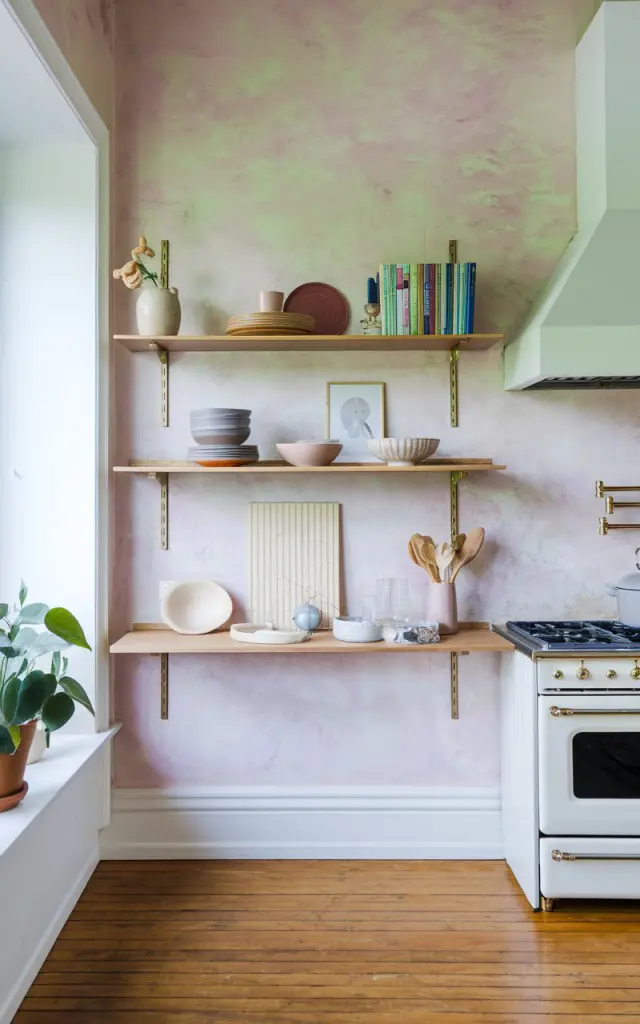 A photo of a minimalistic boho kitchen with sleek, modern floating open shelves. The shelves hold dishes, books, and decor. The wall has a textured pastel color. The kitchen has a wooden floor and a white range hood. There is a potted plant near the window.