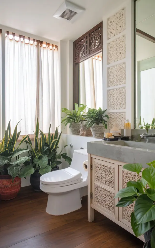 A photo of a Balinese chic bathroom with a toilet, sink, and vanity. There are green plants (snake plants, pothos) placed strategically around the room. The bathroom has a ventilation system and light filtered through curtained windows. The walls are adorned with intricate carvings. The floor is made of wood.