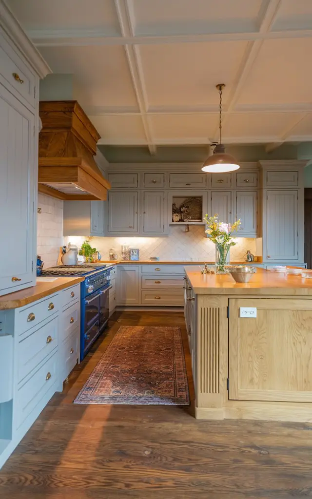 A photo of a honey oak kitchen with light grey cabinets, a honey oak island, and rangehood. The kitchen has neutral walnut stain flooring and a contrasting rug. The kitchen and island are fully furnished. There is a suspended ceiling with a pendant light. The evening light is ambient and warm.