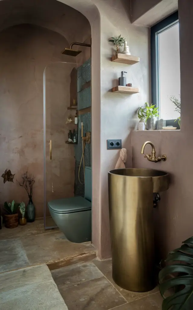 A photo of a small, modern rustic bathroom with a muted color palette. In the corner, there's a sleek, pedestal sink with a brass faucet. The sink is positioned next to a window. The floor is made of large, beige tiles. There's a shower area with a glass door next to the sink. The wall above the sink has a wooden shelf with a few items. There's a toilet in the bathroom. The room has a few plants and decorative items. The lighting is soft.