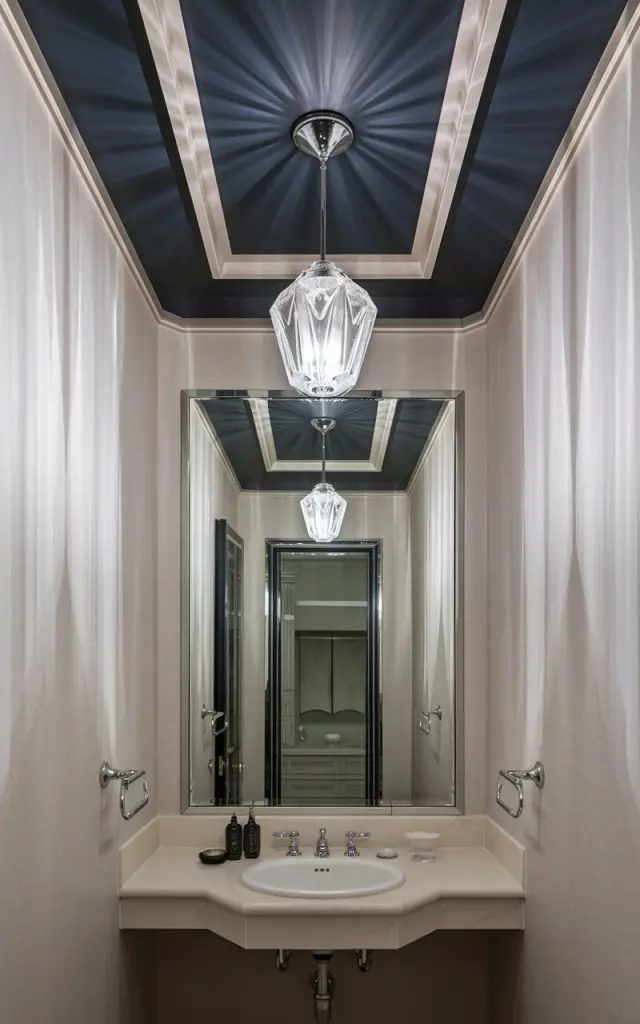 An art deco powder room with light-colored walls and a dark-colored (navy and charcoal) recessed ceiling. There is a glass crystal pendant light hanging from the ceiling. The room is fully furnished with a sink, mirror, and other bathroom fixtures. The natural light is filtered, creating a soft glow.