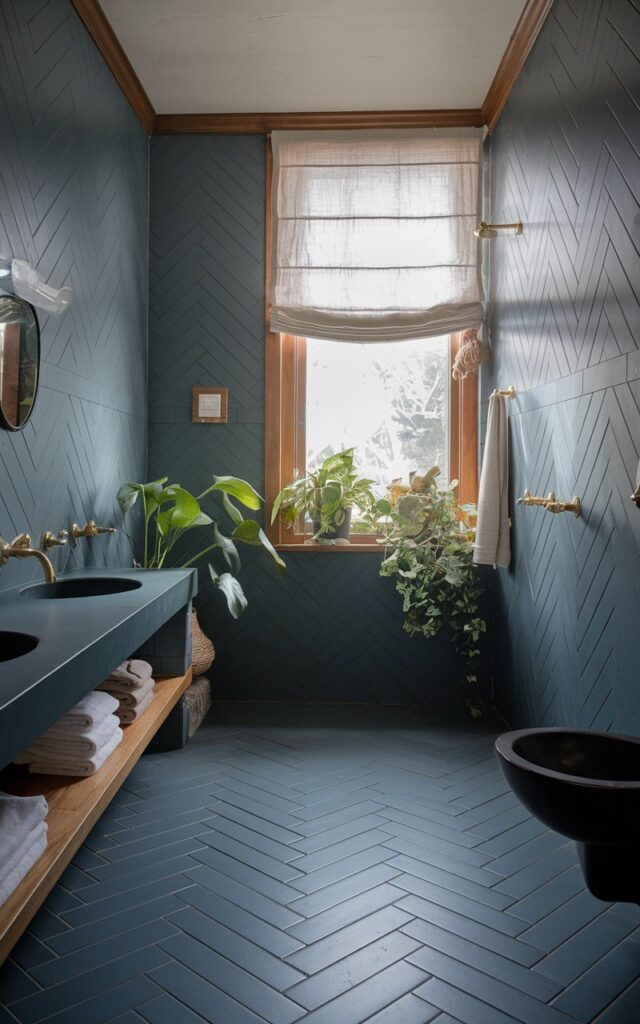 A photo of a California coastal-style moody bathroom with floor-to-ceiling matte slate-blue tiles in a herringbone pattern. The room has a few plants, warm wood accents, and brushed gold fixtures. Soft natural light filters through a gauzy linen curtain window, adding a breezy yet dramatic feel. The bathroom is fully furnished and has a serene and textured atmosphere.