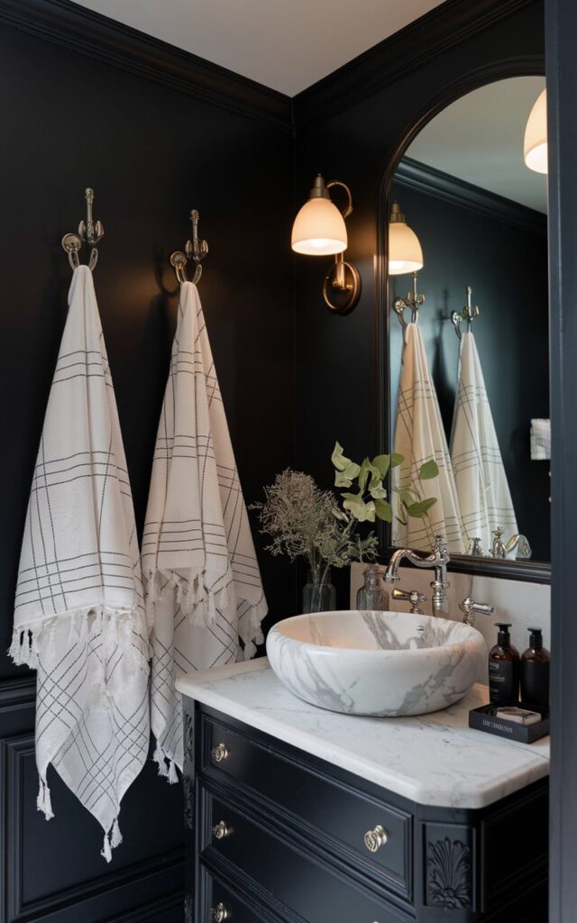 A photo of a French country bathroom with black walls, featuring 2-3 white designer towels hanging on hooks. The vanity has a white marble veined vessel sink with essentials. The vanity has dimmable lights. The bathroom is fully furnished and well-decorated.
