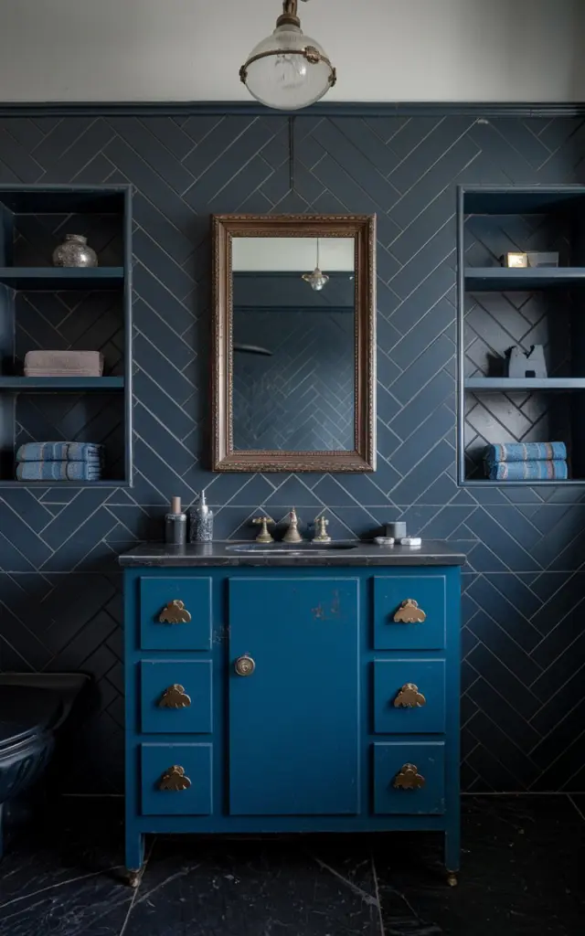 A photo of a Gustavian moody bathroom with a vintage deep blue vanity. The vanity has small brass cloud-shaped knobs and a framed mirror above it. The walls have dark tiles in a chevron pattern and open shelves for decor and towels. The floor is made of dark marble. The ceiling has a vintage-style pendant light that is turned on. The bathroom is fully furnished. The focus is on the vintage vanity and brass knobs.