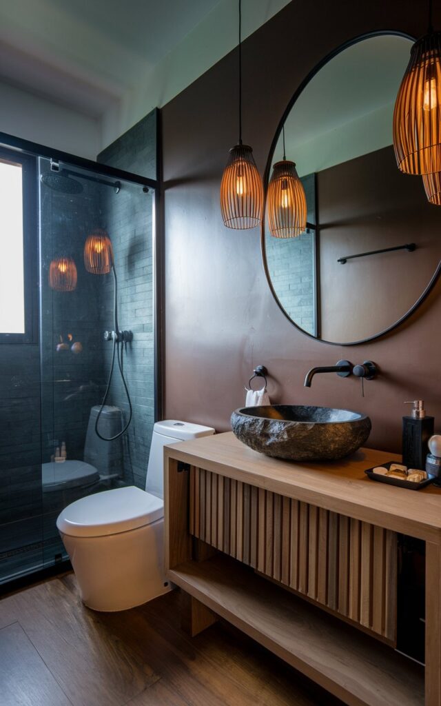 A photo of a modern Balinese bathroom with a moody hue. The bathroom has a vanity with a charcoal stone vessel sink and a black faucet. The bathroom is fully furnished, with a toilet, a shower, and a mirror. There are pendant lights with warm light above the vanity. The floor is made of wood.