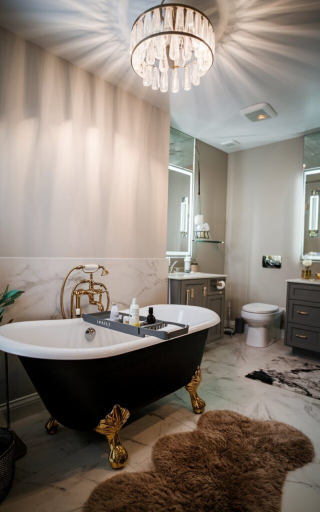 A photo of a modern English countryside style bathroom with a black freestanding tub with golden legs. There is a bathtub caddy with essentials placed on the tub. The ceiling has a chandelier that is lighted. The floor has a fluffy rug. The bathroom also contains a toilet, vanity, and other fixtures. The natural light is soft and ambient.