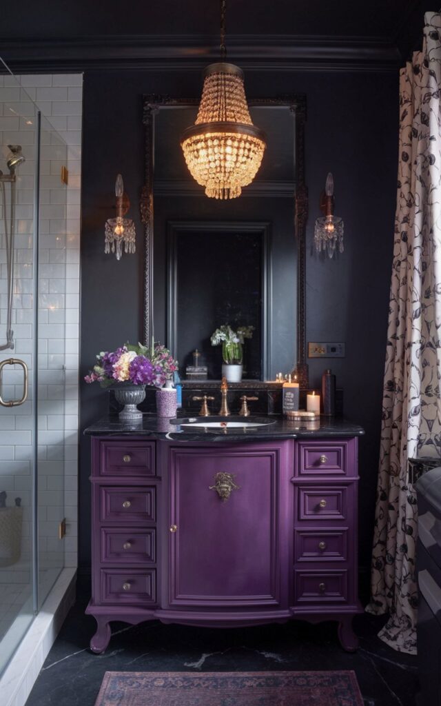 A photo of a moody French country bathroom with an eggplant-colored vanity. The vanity has a rich satin finish and is topped with black marble. It features brushed gold hardware. The deep purple hue adds a luxurious, unexpected pop against the charcoal walls. A vintage chandelier hangs overhead, casting a warm bright glow. The vanity is furnished with essentials, decor like a flower vase, and candles. The bathroom has a shower area and a floor rug.