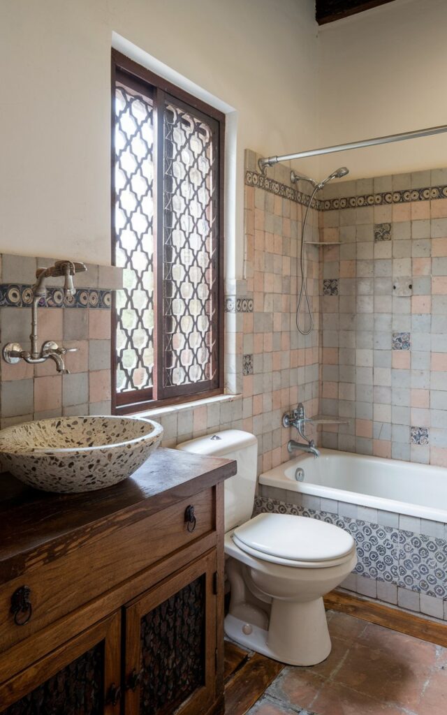 A photo of a south traditional bathroom. There's a terrazzo vessel sink on a wooden vanity. The bathroom is fully furnished with a toilet, a bathtub, and a shower. There's a window with a decorative grille. The walls are adorned with decorative tiles. The floor is made of ceramic tiles.