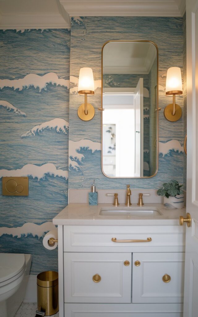 A photo of a transitional + chic half bathroom with a coastal-theme wallpaper and gold accents. The wall has a pattern of blue waves with white foam at the top. There are two gold wall scones on either side of a mirror above the vanity. The vanity has a white countertop and a gold faucet. There is a toilet in the background. The room has a gold trash can and a towel bar. The light is warm and soft.