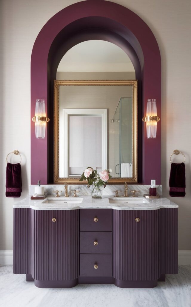 A photo of a regal and chic bathroom with a luxurious yet modern aesthetic. The room features a sleek double-sink vanity with fluted cabinetry and a large painted arch rising from the countertop to the ceiling. The arch is painted in a deep plum hue, contrasting against the soft neutral walls. Above the arch, a gold-framed mirror and matching wall sconces (warm light on) add sparkle and symmetry. The countertop is made of marble, and the hardware is brass. Velvet-textured hand towels and a small crystal vase with fresh flowers complete the elegant, fashion-forward atmosphere. Natural light is soft, ambient.