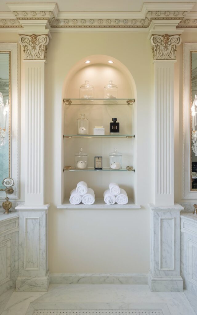 A photo of a fully furnished bathroom with a regal white theme. The bathroom has a built-in wall niche painted the same shade of soft white as the surrounding walls. Inside the niche, there are glass jars, rolled towels, and perfume bottles. The niche is lit with subtle recessed lighting. The bathroom has ornate moldings, gold accents, and marble details.