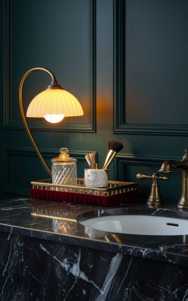 A photo of a Victorian + modern fusion bathroom vanity with a striking black marble countertop and brass fixtures, set against a panelled wall in deep green or navy. There is a clip-on small, stylish lamp with a curved brass neck and a milk-glass or pleated shade, on the edge of the vanity, glowing softly with warm yellow light. The lamp casts a cozy ambiance over the counter, which holds a mix of vintage and modern elements—cut-glass jars, a velvet-trimmed tray, and a sleek gold makeup brush holder. The blend of ornate details and clean lines creates a space that’s moody, refined, and perfectly layered. No natural light.