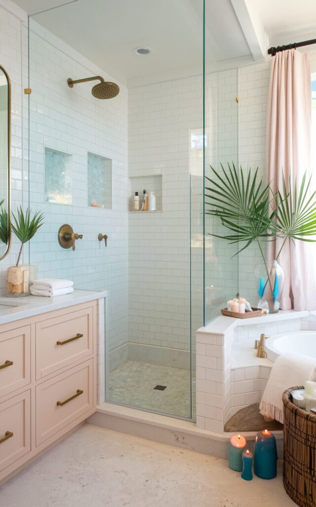 A photo of a bright and breezy California Coastal meets Palm Beach bathroom. The shower is defined by a sleek frameless glass partition. The walls are covered in crisp white subway tiles with soft sea-glass accents. The flooring features pale sand-toned stone. There's a brass rainfall shower head and recessed shelves with essentials. The room has a blush-toned vanity with gold hardware. There are coastal textures like linen, cane baskets, and driftwood accessories. Palm fronds sit in a glass vase near the curtained window, while ocean-scented candles rest on the ledge beside the tub.