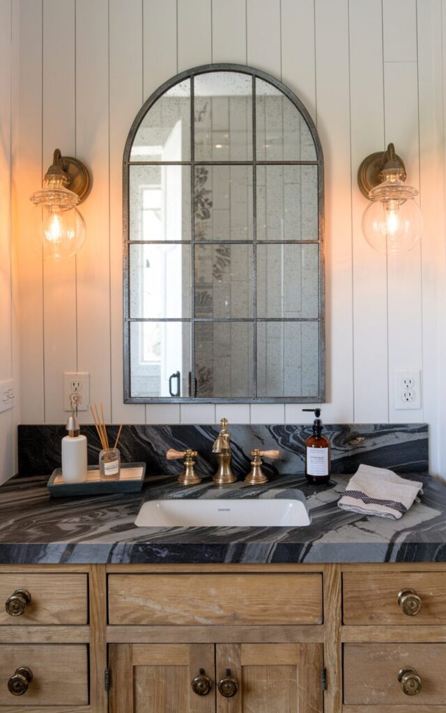 A closer view of the marble slab countertop in the farmhouse-luxe bathroom. The marble is rich with bold gray and charcoal veining, creating a striking contrast against the raw wood tones. The vintage-style gold faucets and matching drawer pulls add a glamorous touch. The oversized arched mirror with a distressed metal frame hangs above, reflecting the room. The walls are covered in white vertical shiplap, and sconces with clear glass globes bring warm ambient light. A ceramic tray with perfume bottles and a linen hand towel complete the elevated yet grounded vibe.