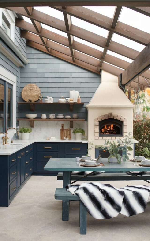 A photo of a fully furnished alpine chic outdoor kitchen attached to a house. The kitchen has navy blue cabinets, brushed brass hardware, white marble countertops, and a stone backsplash. There's a wood-fired oven, open shelving with ceramic dishware, and a large farmhouse dining table with faux fur throws draped over benches. The space is under a sloped pergola roof supported by exposed timber beams. The ambiance is cozy yet fresh, blending alpine warmth with coastal elegance.
