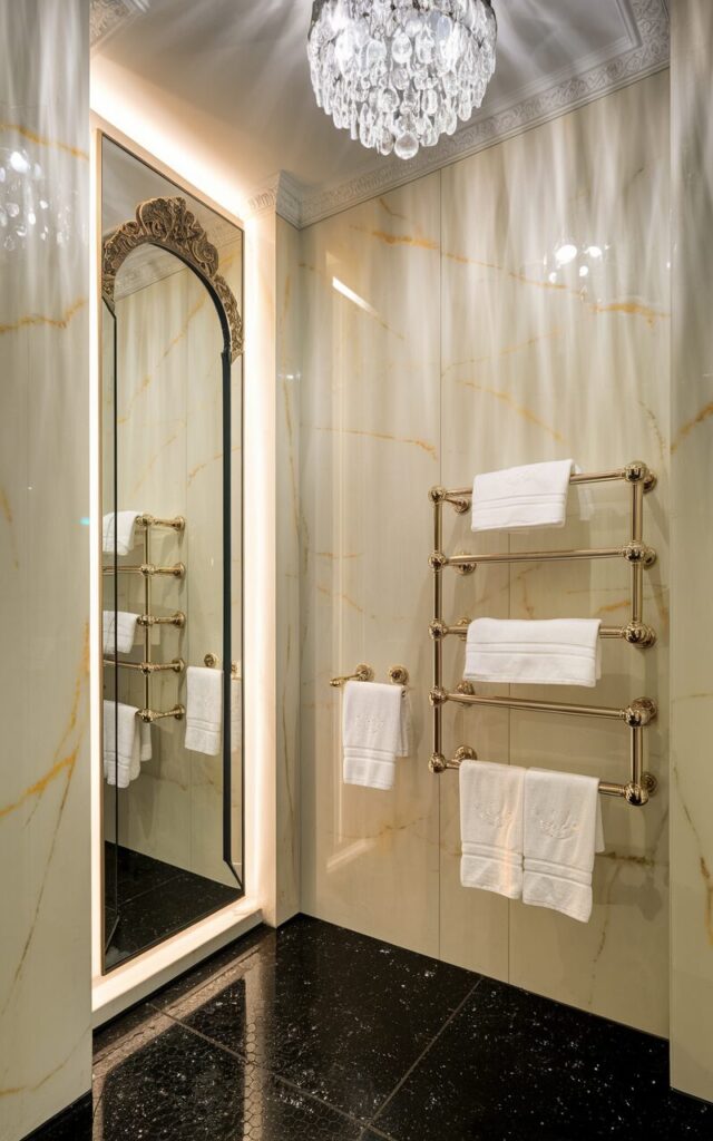 A photo of a glamorous bathroom with a sleek, modern vibe. The walls are made of soft marble with subtle gold veining. There is a large ornate mirror with backlighting. Elegant floating towel bars in polished brass are mounted on the wall, holding plush white towels neatly folded. The floor sparkles with black tiles. A crystal chandelier hangs from the ceiling, adding sparkle from above.