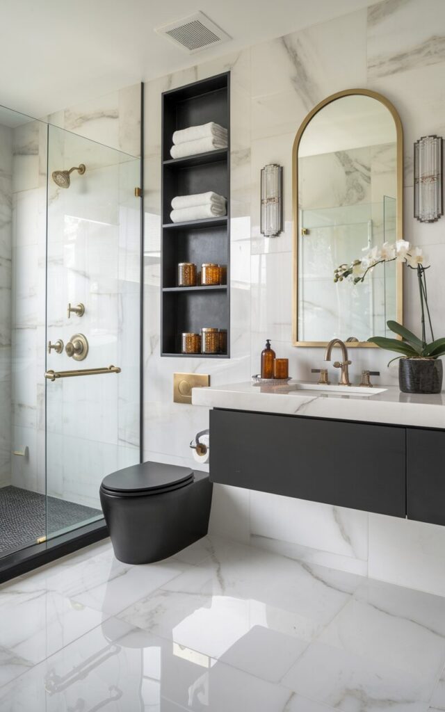 A photo of a glamorous full bathroom with a seamless, luxurious feel. The large-format marble tiles on both the floor and walls have a white base with mild gray veining. Contrasting matte black hardware, including a sleek wall-mounted faucet, a brass towel bar, and brass shower fittings, pop against the glossy marble. A modern back-to-wall toilet with a matte finish sits nearby, framed by a vertical wooden shelf holding folded plush towels, gold-rimmed jars, and a scented candle. Above the floating vanity, a bold arched mirror with a gold frame anchors the space, flanked by crystal-like wall sconces. The decor includes a potted orchid, amber glass accessories, and a sculptural soap dish. The entire bathroom is polished, high-contrast, and ultra-chic.