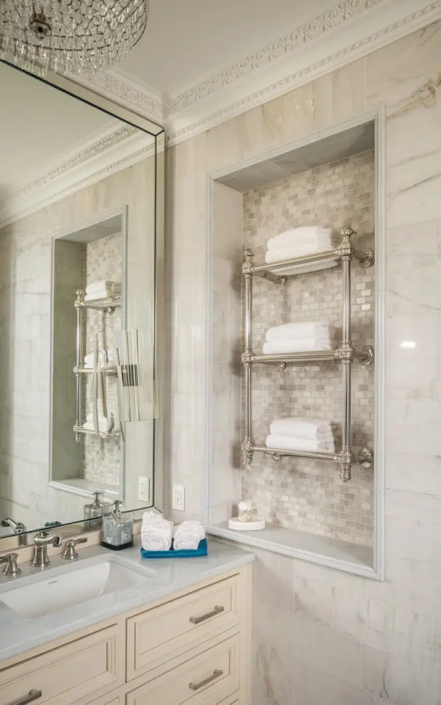 A photo of a glamorous-style bathroom with soft marble walls, brushed nickel fixtures, and elegant crown molding. The room has a recessed niche lined with subtle mosaic tiles and houses sleek polished towel bars with plush, neatly folded towels. The niche is framed with delicate trim, blending seamlessly into the space. A sparkling chandelier hangs above, and a large framed mirror reflects the light, adding a sophisticated yet approachable charm to the room.