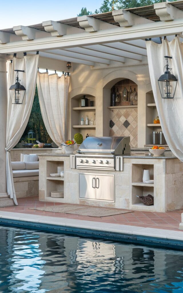 A photo of a luxurious European-style outdoor kitchen beside a shimmering swimming pool. The kitchen has a built-in stainless steel grill station with elegant stone cladding. The grill is centrally positioned with ample counter space on both sides for prep and serving, topped with polished granite. Classic European details like wrought iron lanterns and terracotta tiles frame the space. A pergola draped with white linen curtains surrounds the kitchen, providing seating and a dining area. The kitchen has other functional elements such as open shelves with essentials and a floor rug. The swimming pool is in the backdrop.