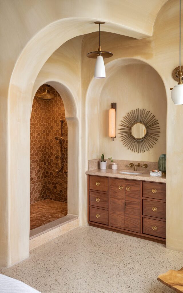 A photo of a mid-century modern and Mediterranean ensuite bathroom with graceful archways. The room has a wide, rounded arch connecting the bedroom to the ensuite. Inside, smaller arches define zones: one leads to the walk-in shower with zellige tiles in warm clay tones, and another to a built-in vanity alcove with walnut cabinetry and brass pulls. The floors are terrazzo or travertine, and the walls are creamy stucco. Mid-century pendant lights and a sunburst mirror add retro charm to the room.