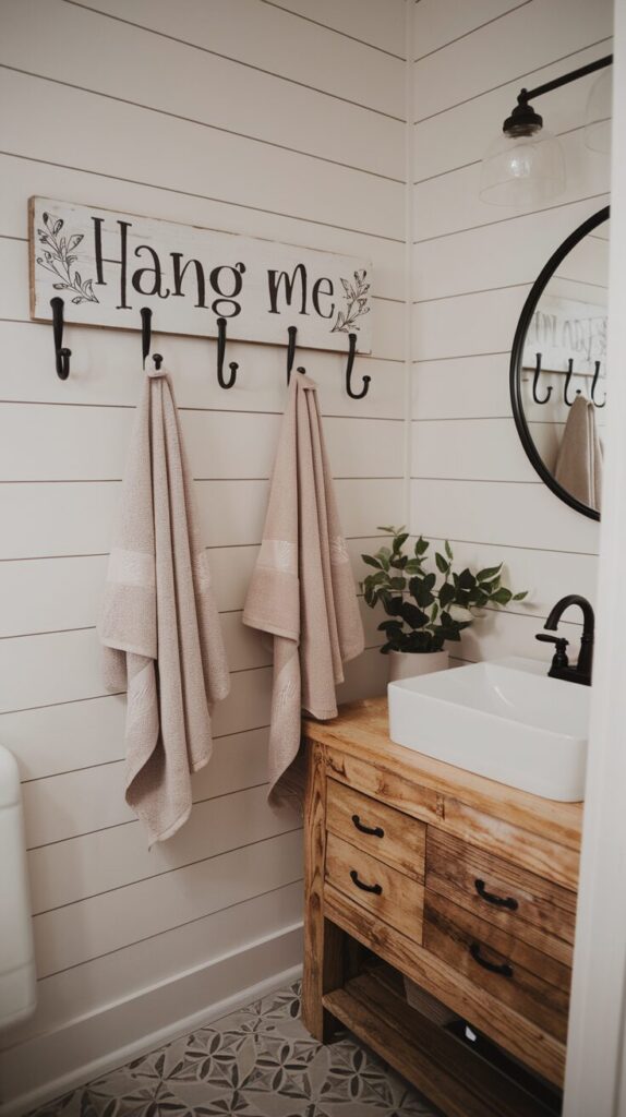A photo of a modern farmhouse bathroom with shiplap walls painted crisp white, matte black fixtures, and warm natural wood accents. A rustic wooden plank with the handwritten line "hang me" in black Italic font with floral designs is mounted horizontally on the wall. The plank has evenly spaced black iron hooks. Soft, neutral-toned towels are hung on the hooks. Below the plank, a white porcelain sink sits on a reclaimed wood vanity. A round black-framed mirror hangs above the sink. The floor features patterned tiles in muted gray tones. A small potted plant adds a cozy, fresh touch to the space.