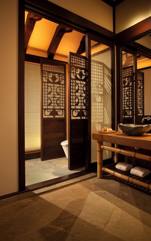 A photo of a serene ensuite bathroom with a Japanese-inspired design. The bathroom is connected to the bedroom and features a toilet hidden behind a dark wooden screen with intricate stencil patterns. The screen casts soft shadows and filters light. The bathroom has a wooden vanity with a vessel sink and bamboo accents. The floor is made of natural stone. The lighting is warm amber. The overall atmosphere is tranquil and luxurious.