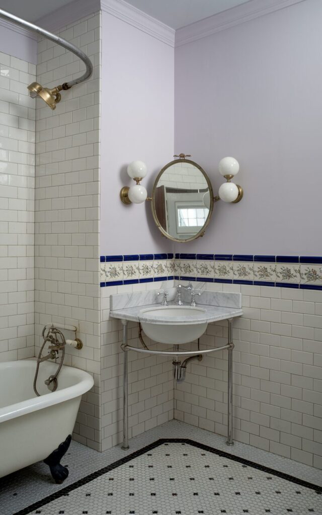 A photo of a Victorian-style bathroom with a corner sink mounted on a marble-topped vanity. The room has white subway tiles on the walls and a decorative floral border tile. Above the sink, a petite oval mirror with a vintage brass frame is flanked by twin globe sconces. The room also contains a clawfoot tub with a curved shower curtain rail and black-and-white mosaic floor tiles. The walls are painted in soft, pale lavender.