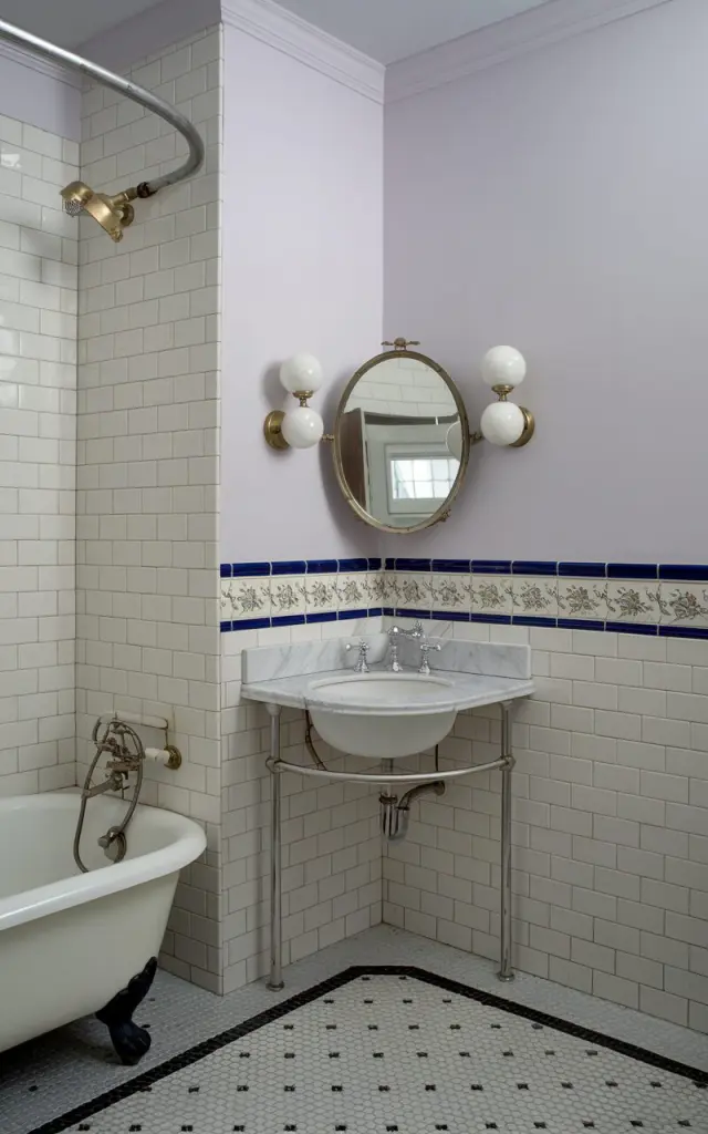 A photo of a Victorian-style bathroom with a corner sink mounted on a marble-topped vanity. The room has white subway tiles on the walls and a decorative floral border tile. Above the sink, a petite oval mirror with a vintage brass frame is flanked by twin globe sconces. The room also contains a clawfoot tub with a curved shower curtain rail and black-and-white mosaic floor tiles. The walls are painted in soft, pale lavender.
